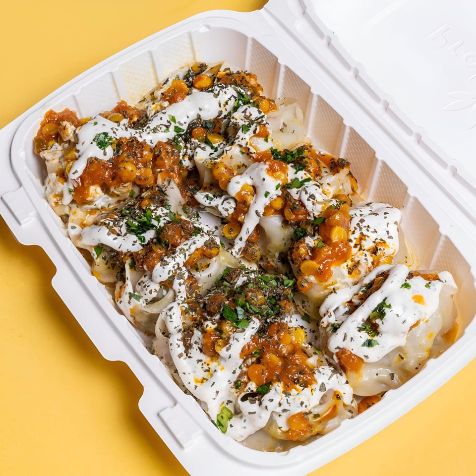 Sheesh Grill's middle-eastern dumplings topped with white sauce, red sauce, and herbs in a takeout container.