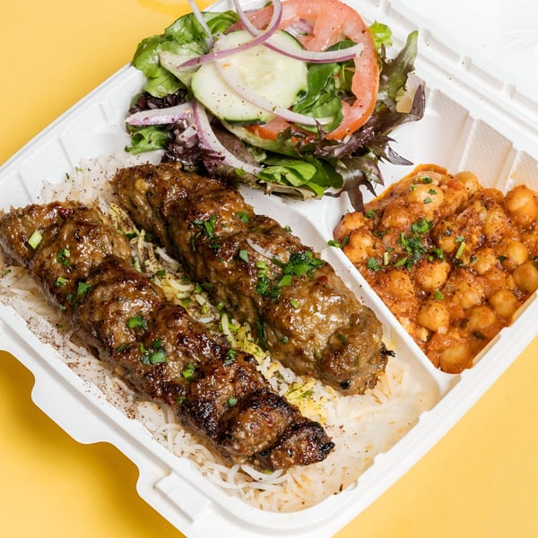 Two grilled Adana kebabs on rice, with a side salad and chickpeas in a takeout container.