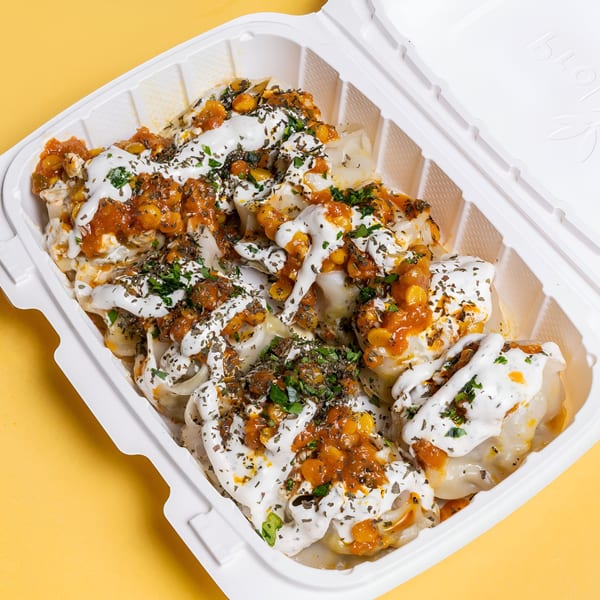 Middle-Eastern dumplings topped with white sauce, red sauce, corn, and herbs in a white takeout container.